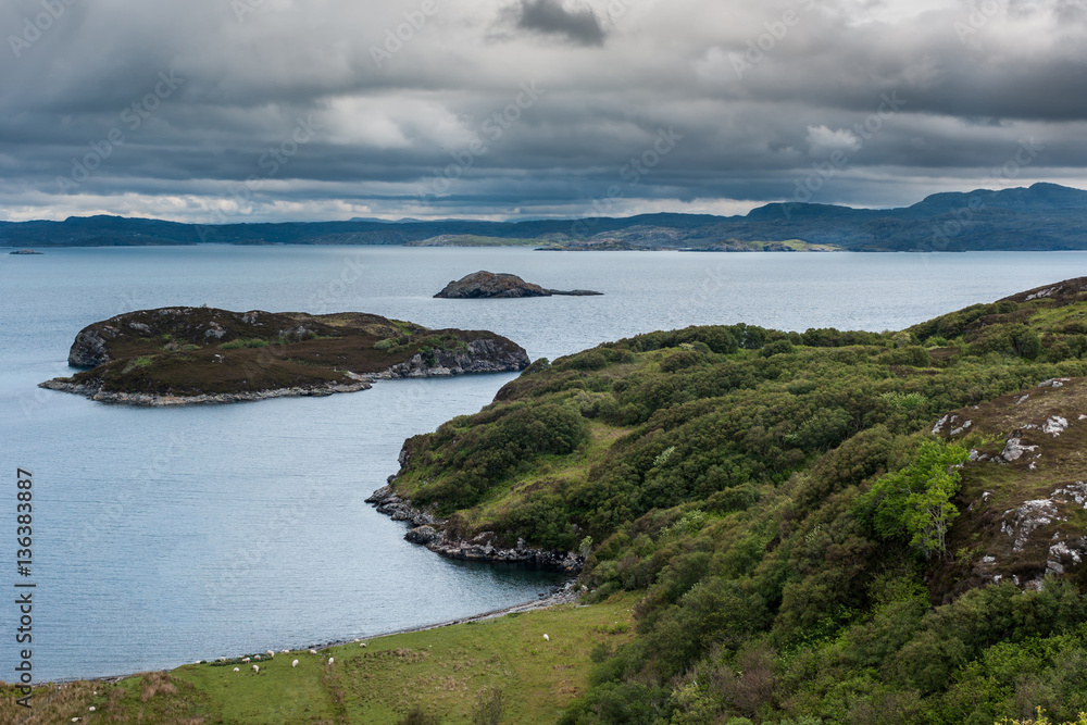 Assynt Peninsula, Scotland - June 7, 2012: View from higher up Drumbeg hamlet onto Atlantic Ocean under dark cloudy sky. Rocky islands with green patches, gray seas, and some vegetation upfront.