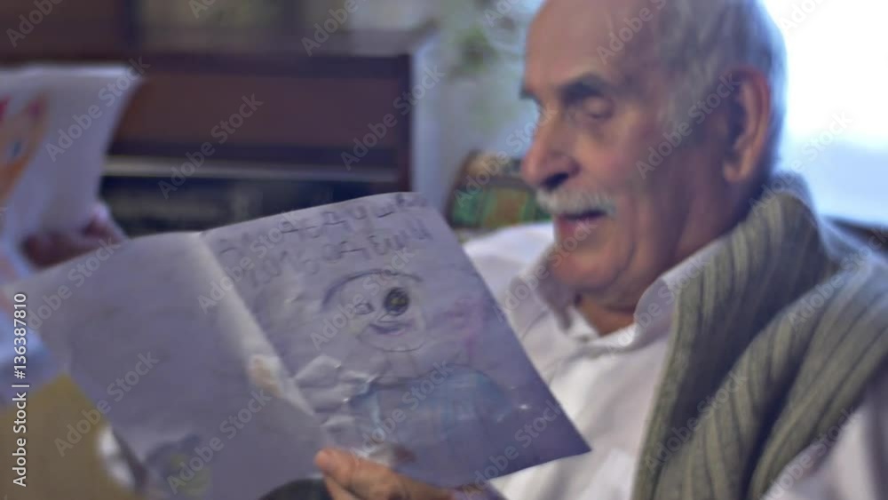 Senior Man is Glad to Receive a Gift From Kids Holding the Drawings and Shows it Proud of His Happy United Family Grandad is Not Lonely in Cosy Room Senior Man is Glad to Receive a Gift From Kids Holding the Drawings and Shows it Proud of His Happy United Family Grandad is Not Lonely in Cosy Room