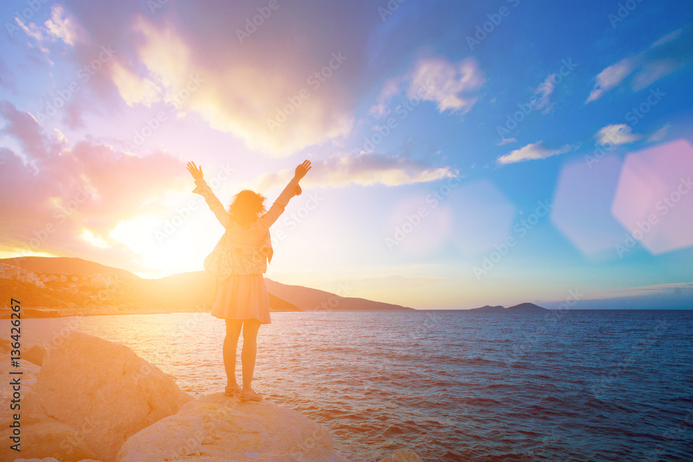 Woman with her hands raised standing on a rock near the sea at sunset
