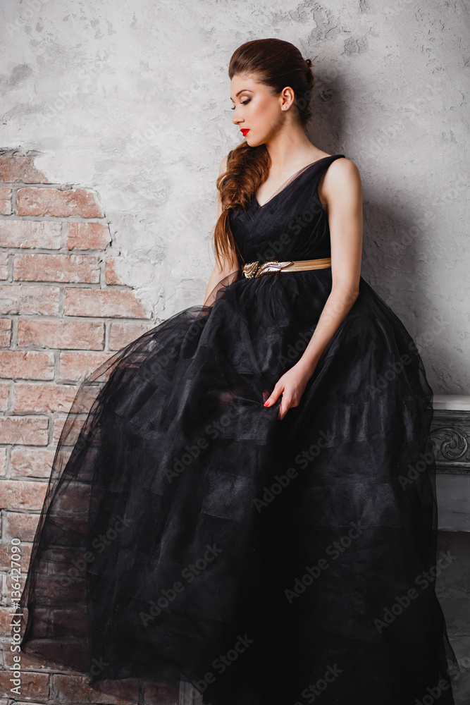 beautiful girl with bright makeup sitting on the fireplace in a long, black dress shoes with pigtail looking down on a light brick background