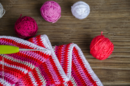 crocheted striped fabric in red colors with crochet hook and many small yarn balls on the old wood background