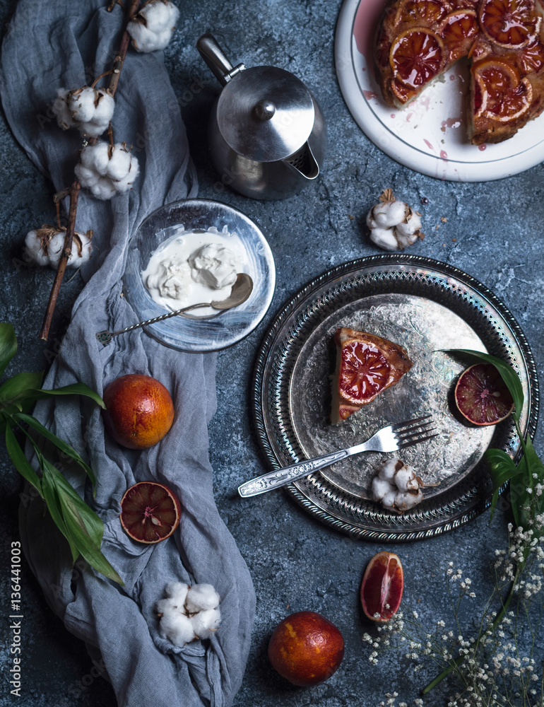 blood orange upside down cake on a white stand with a slice on a dish with oranges, cotton, green leaves, ice cream and a teapot on a blue background blood orange upside down cake on a white stand with a slice on a dish with oranges, cotton, green leaves, ice cream and a teapot on a blue background