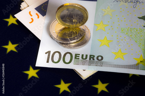 European cash money. Several 50 euro bills, 100 euro bill above them. Three euro coins on the hundred euro bill. One coin is standing. European community flag as a background. Macro image. photo