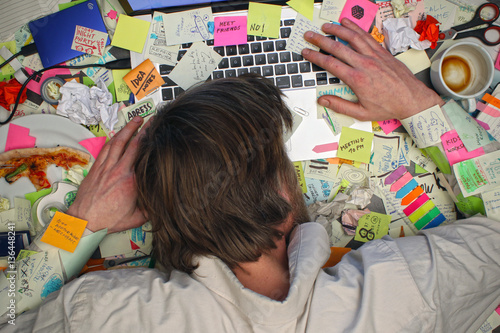 Overworked man sleeping on laptop computer with post it notes all around his office desk. photo