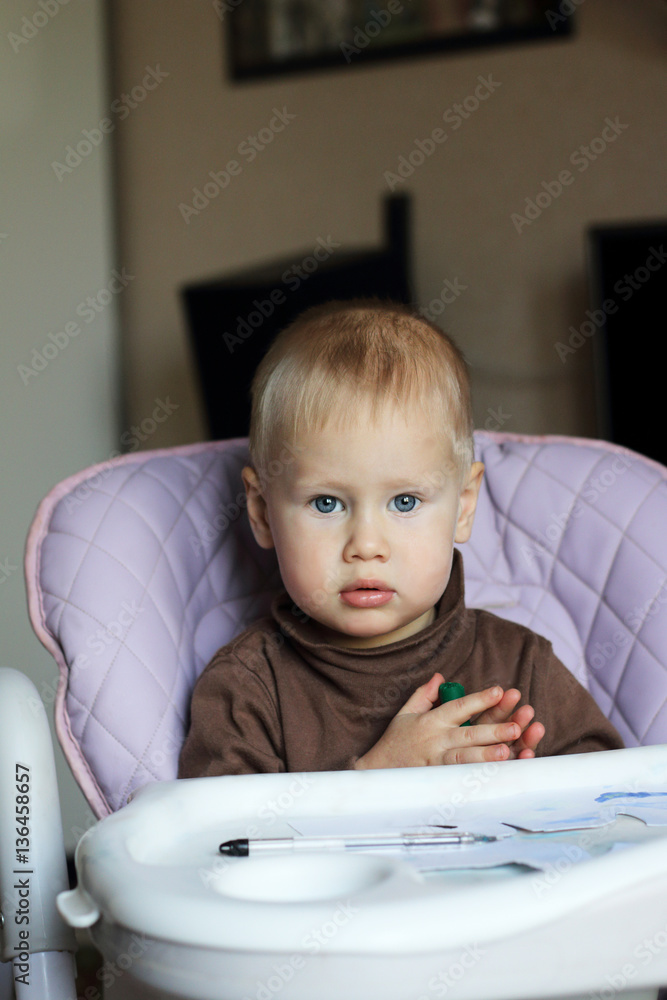 Little blonde ordinary baby boy sitting at chair for feeding and draws Little blonde ordinary baby boy sitting at chair for feeding and draws