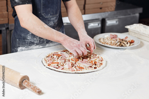 Cooking pizza. arranges meat ingredients on the dough preform. Closeup hand of chef baker in uniform blue apron cook at kitchen