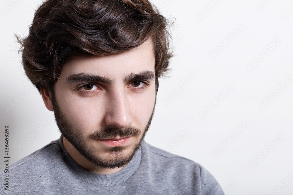 Fototapeta premium Portrait of sullen angry man with trendy hairstyle and dark beard wearing grey casual sweater looking at camera with serious and severe face expression, demonstrating his masculanity.