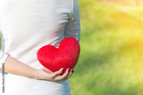 Fototapeta Naklejka Na Ścianę i Meble -  Asian woman holding red soft heart pillow with one hand near her waist at park as background to show her love on Valentines day with warm yellow sun light