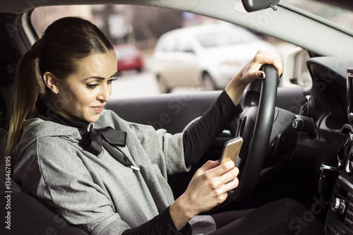 Woman texting while driving. Beautiful blonde woman driving her automobile and texting on her smartphone.