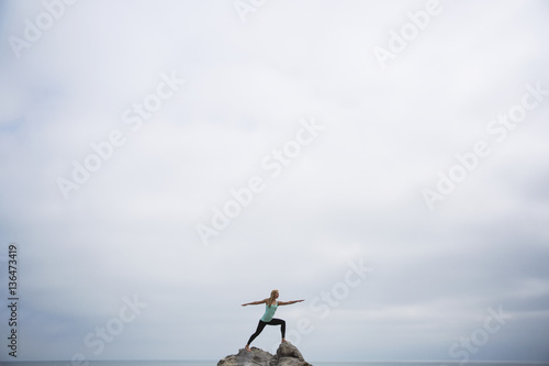 Woman practicing worrier 2 pose on rocks in sea against cloudy sky