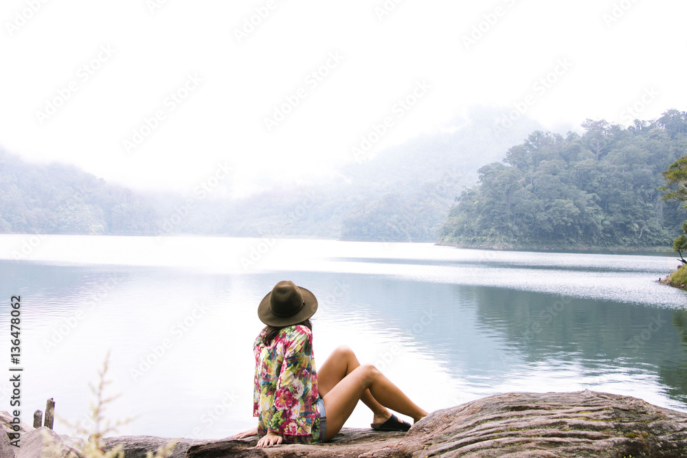 Side view of woman in hat sitting on log by lake during foggy weather Side view of woman in hat sitting on log by lake during foggy weather