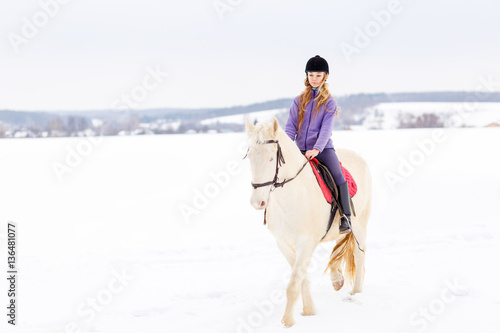 Young girl in helmet riding white horse on winter field