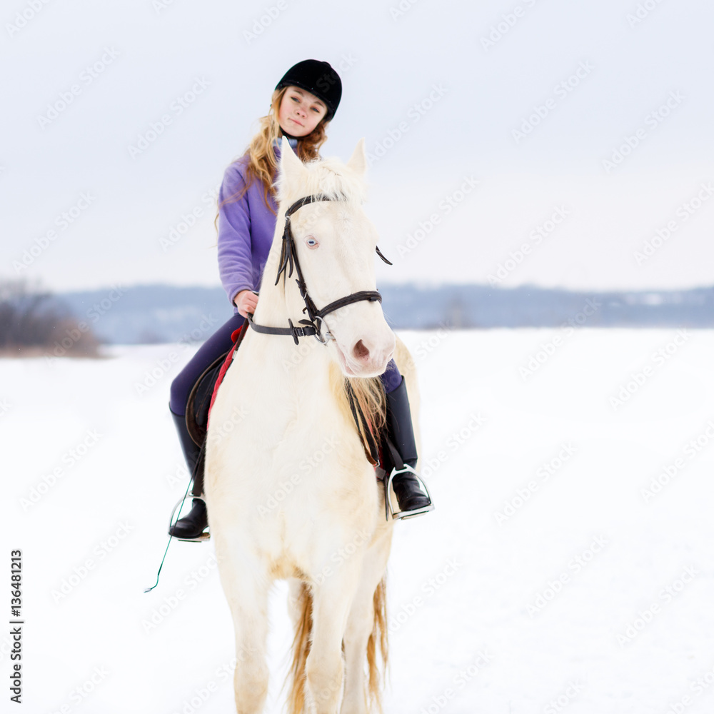 Fototapeta premium Young girl in helmet riding white horse on winter field