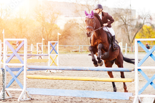Young female rider on bay horse jumping over hurdle on equestrian sport competition