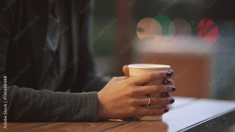 Close-up of woman s hand holding a cup of coffee, fume from the tea cup. Blurred lights, urban street Close-up of woman s hand holding a cup of coffee, fume from the tea cup. Blurred lights, urban street