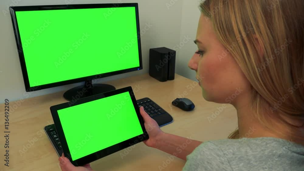 A young woman sits at a desk and looks in turns at a computer screen and a tablet (both green screen) A young woman sits at a desk and looks in turns at a computer screen and a tablet (both green screen)