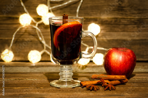 Mug of mulled wine with spices close up. Apple, cinnamon sticks and star anise on a wooden table, lanterns in the background
