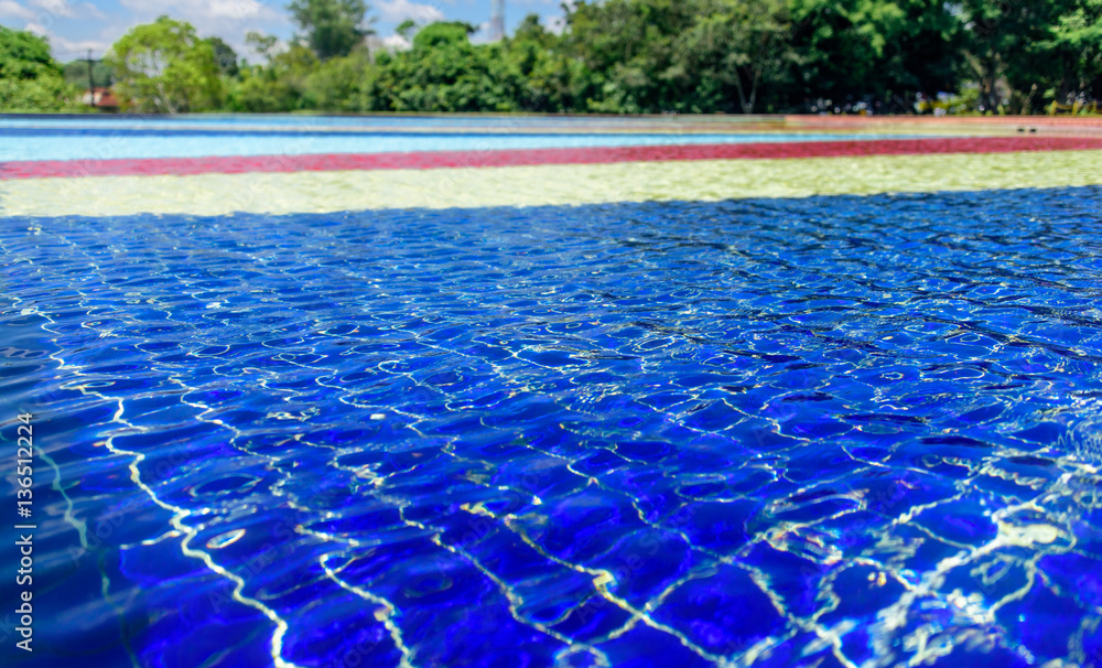 Light blue, red, yellow and blue striped mosaic floor of fountain covered with clear water at sunny day on the background of blury green trees