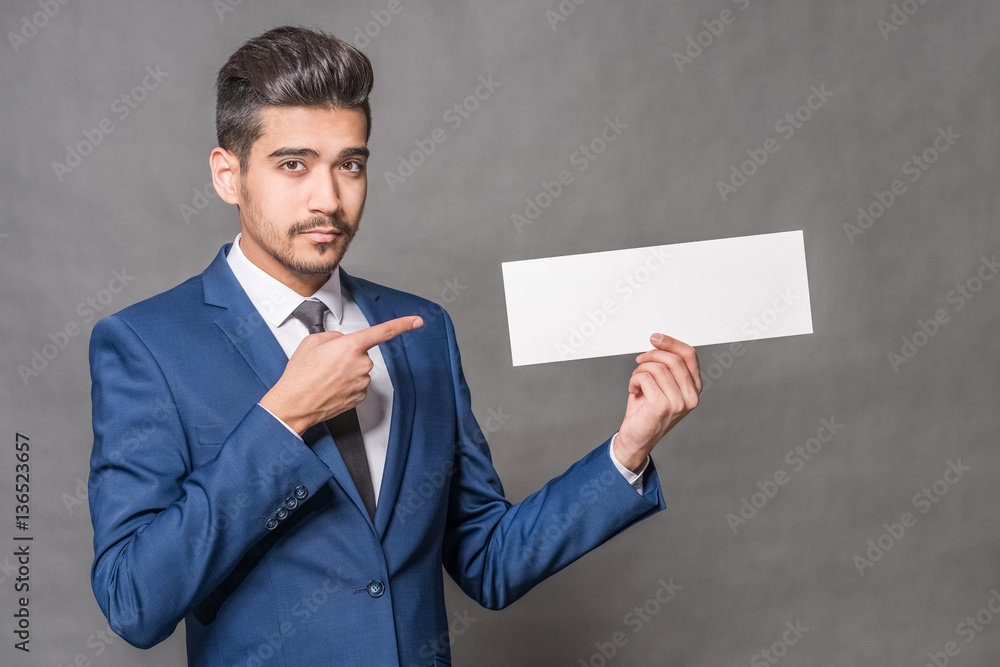 Young attractive man in a blue suit showing with a finger on a empty white sign in hand on a gray background Young attractive man in a blue suit showing with a finger on a empty white sign in hand on a gray background