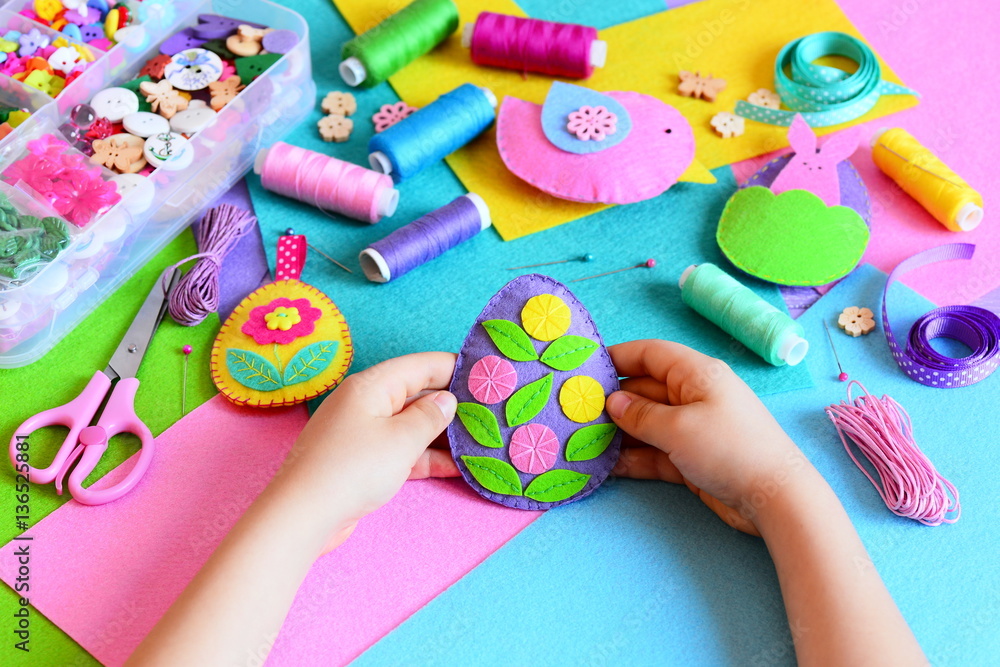 Fototapeta premium Child made a felt Easter egg ornament. Small child holds a felt Easter egg ornament in his hands. Easter crafts, craft tools and materials on a table. Festive spring crafts concept