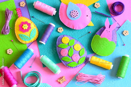 Felt Easter eggs with flowers and bunny, a felt bird. Easter ornaments set, colored thread spools, felt sheets, pins, ribbons, wooden buttons on a table. Colorful Easter background. Top view. Closeup