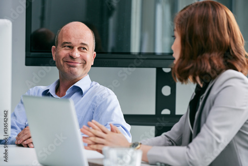 Portrait of smiling middle-aged businessman holding negotiations with his pretty female partner while sitting in meeting room