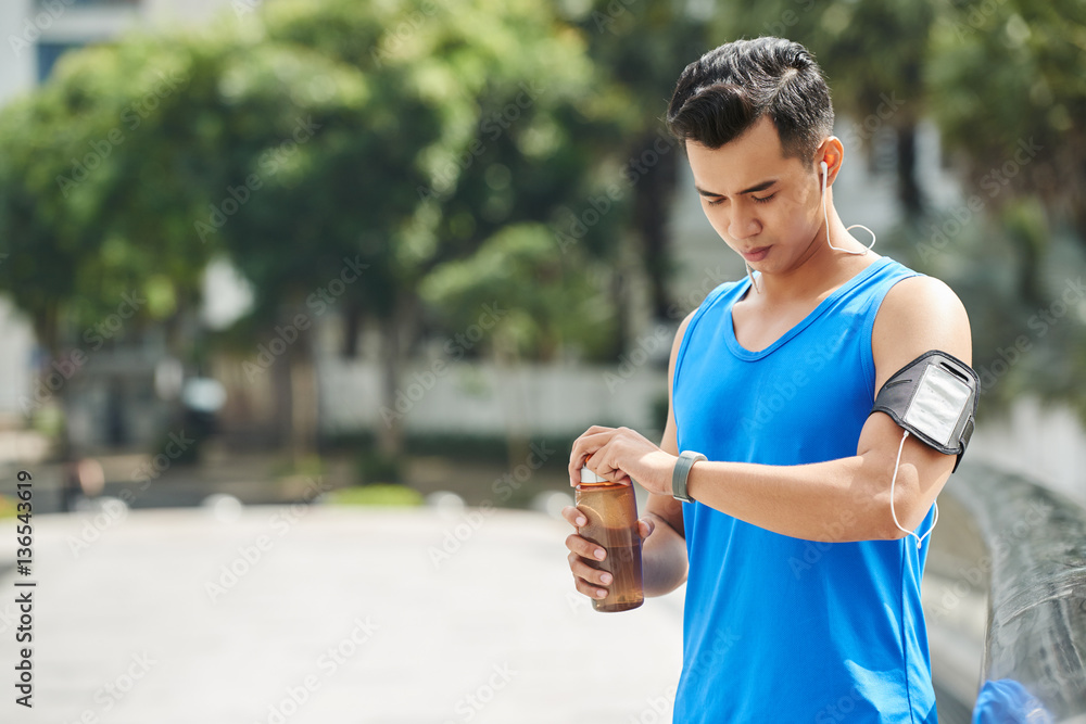 Φωτογραφία Handsome sporty man looking at fitness tracker and opening sports water bottle w