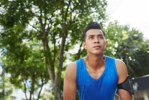 Waist-up portrait of sweaty Asian athlete with smartphone and headphones relaxing after energy-sapping marathon
