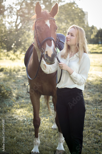 Девушка со светлыми волосами стоит рядом с коричневой лошадью