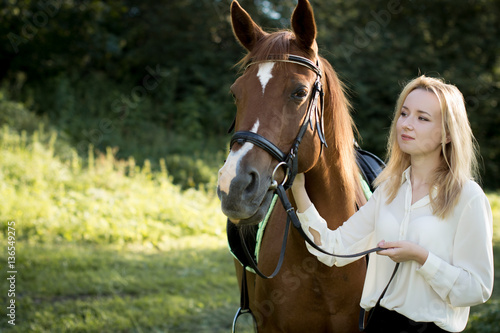 Молодая девушка со светлыми волосами и в белой рубашке стоит рядом с коричневой лошадью в лесу