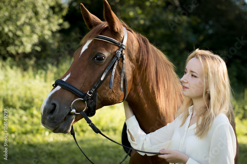 Молодая девушка со светлыми волосами и в белой рубашке стоит рядом с коричневой лошадью в лесу © natasidorova 