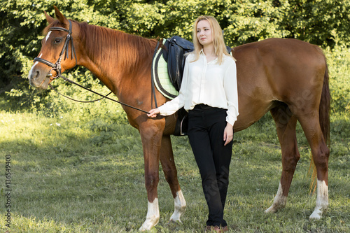 Молодая девушка со светлыми волосами и в белой рубашке стоит рядом с коричневой лошадью в лесу