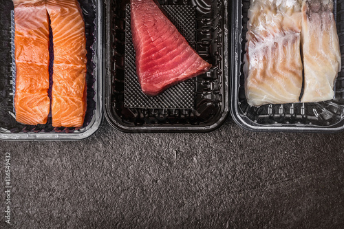 Photography Selection of raw colorful fish fillets : salmon, tuna and codfish in plastic boxes on dark rustic background, top view, border, place for text