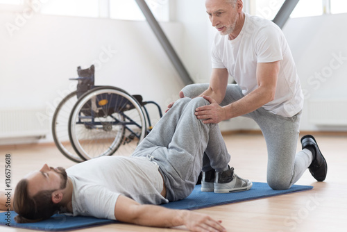 Skilled aged orthopedist stretching the disabled person in the gym photo