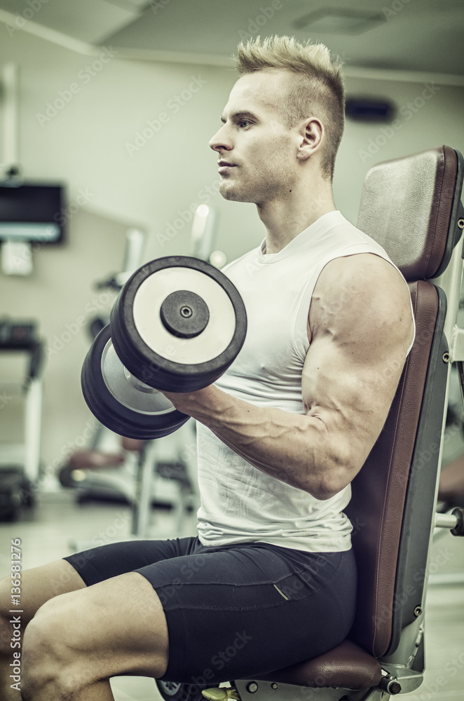 Handsome young man training biceps lifting dumbbells on bench in a gym Handsome young man training biceps lifting dumbbells on bench in a gym