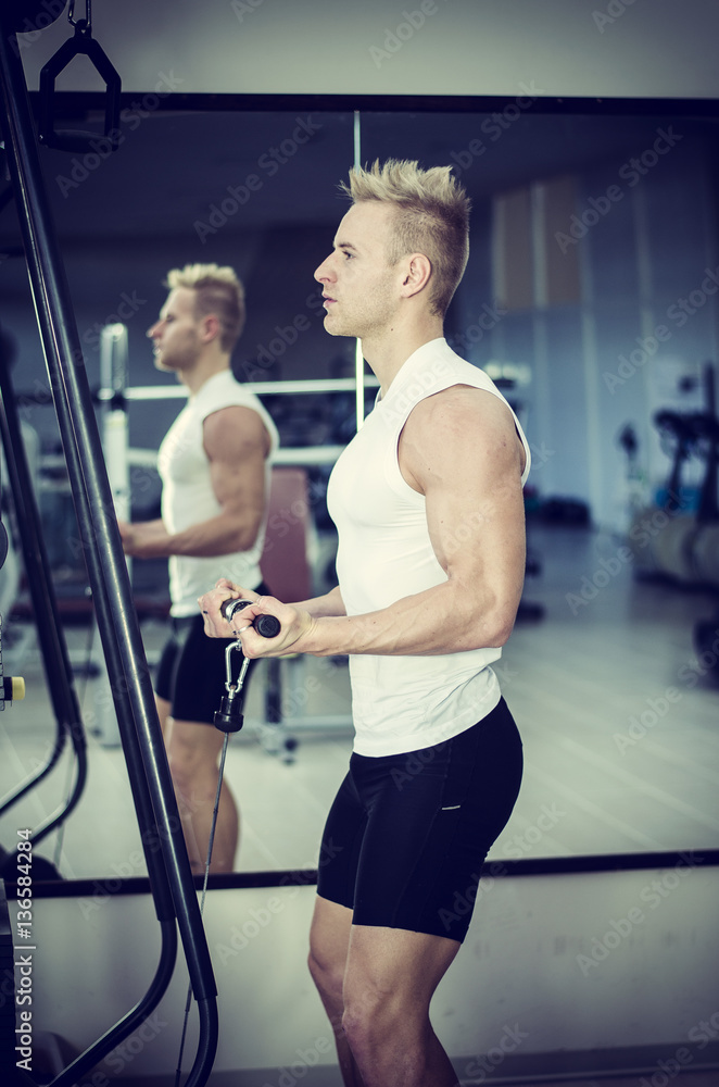 Handsome blond young man exercising triceps on gym equipment, shot from a side Handsome blond young man exercising triceps on gym equipment, shot from a side