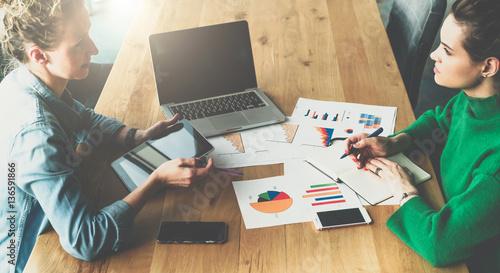 Top view.Business meeting.Two young business women sitting in meeting room at table and discussing.First girl using tablet PC,second takes notes in notebook.On table laptop, smartphones, paper charts. photo