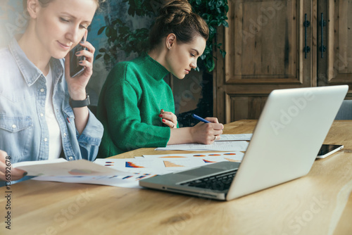 Two young business women sitting in office at table and work together.On table laptop and paper charts.First woman signs documents,second looking charts and talking on phone. Students learning online.