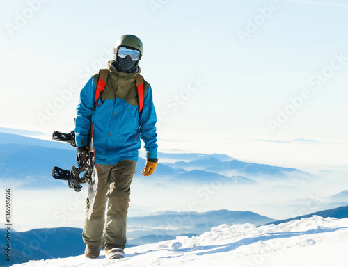 Young snowboarder in helmet walking at the top of a mountain with his snowboard in hand