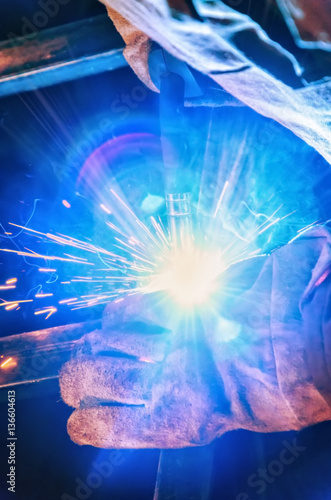 welder in a protective mask in a dark shop floor weld metal parts. By welding sparks fly in different directions