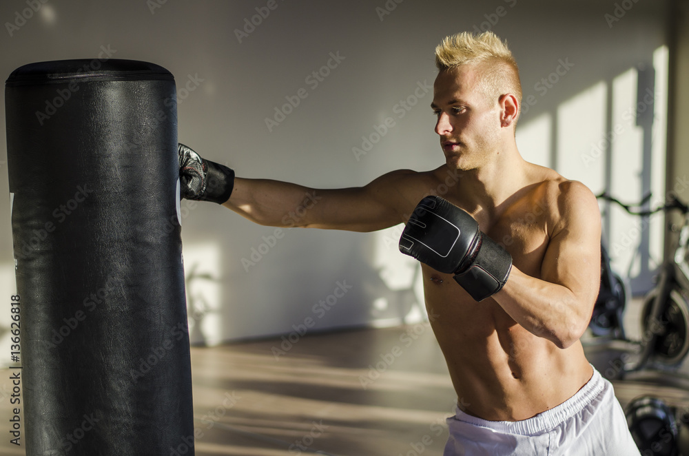 Shirtless handsome muscular young man in gym giving punch to punching bag, wearing boxing gloves