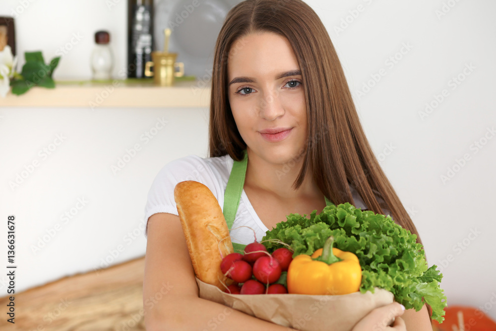 Young caucasian woman in a green apron is holding paper bag full of vegetables and fruits while smiling in kitchen Young caucasian woman in a green apron is holding paper bag full of vegetables and fruits while smiling in kitchen