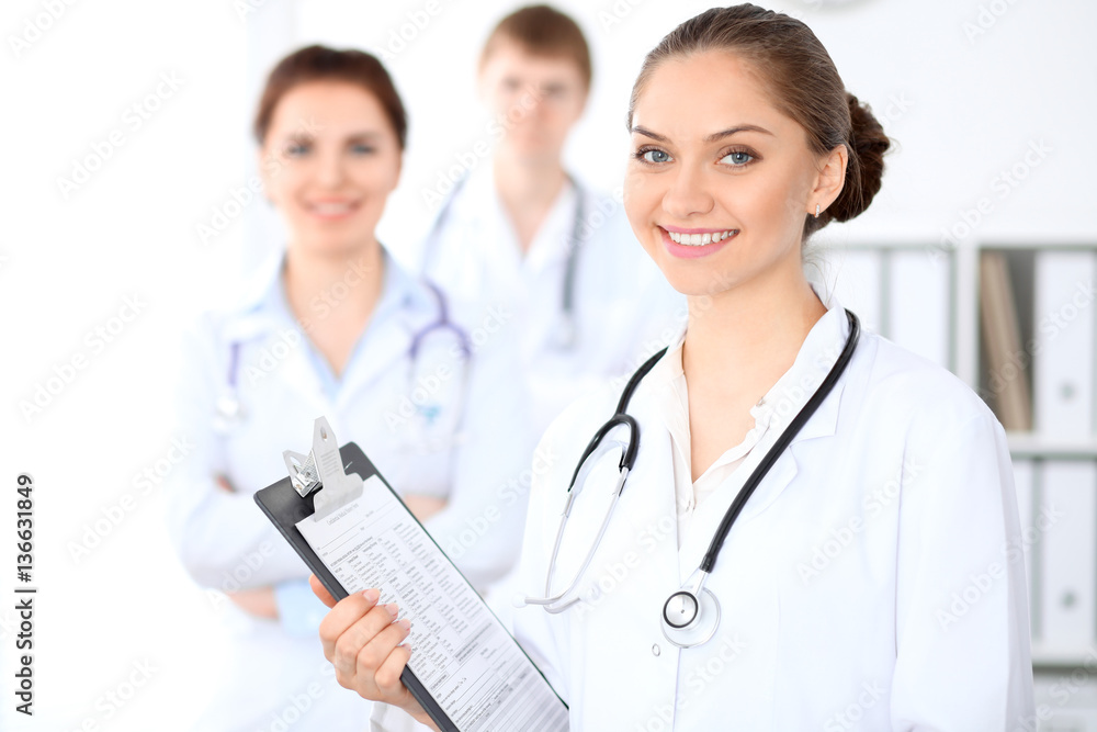 Happy female doctor keeping medical clipboard while medical staff are at the background. Successful team at health care and medicine concept Happy female doctor keeping medical clipboard while medical staff are at the background. Successful team at health care and medicine concept