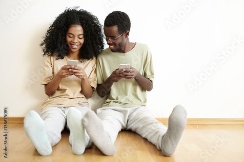 Happy young African married couple having nice time together at home, enjoying free wireless connection, using mobile phones. Man in glasses smiling, looking at screen of his wife's gadget screen