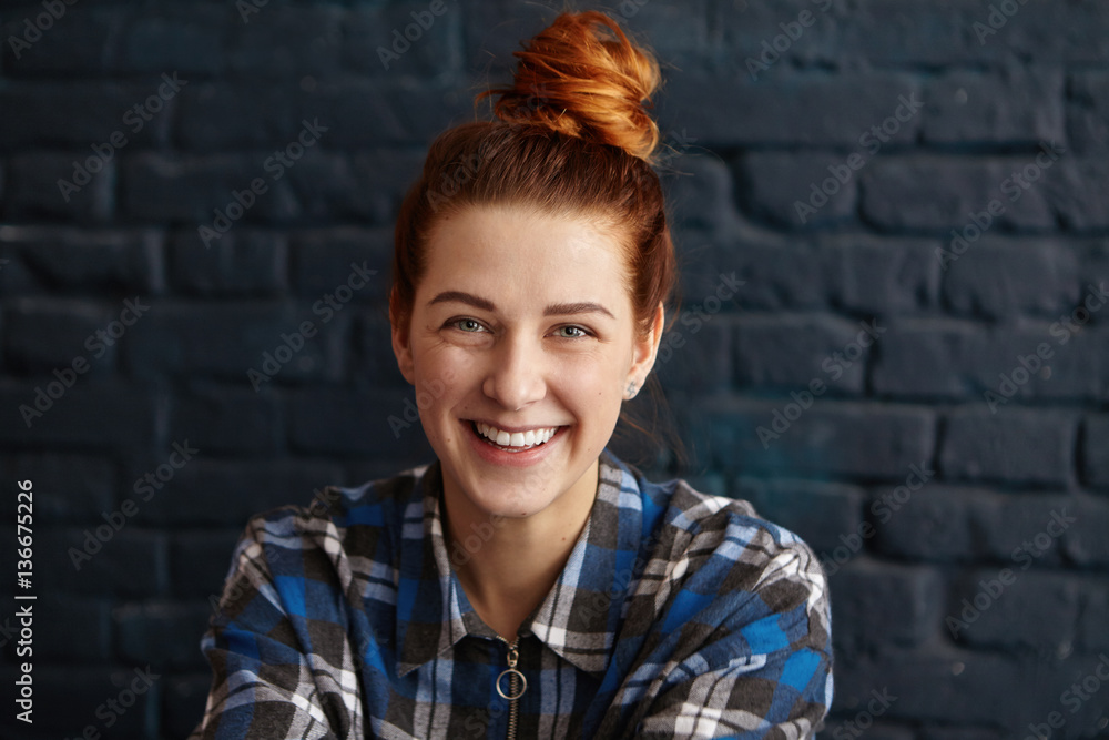 Beautiful young redhead female with hair knot wearing blue checkered shirt looking at camera with cute happy smile, sitting indoors against black brick wall background. People and lifestyle concept Beautiful young redhead female with hair knot wearing blue checkered shirt looking at camera with cute happy smile, sitting indoors against black brick wall background. People and lifestyle concept