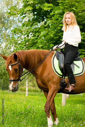 Молодая девушка со светлыми волосами сидит верхом на коричневой лошади в лесу 