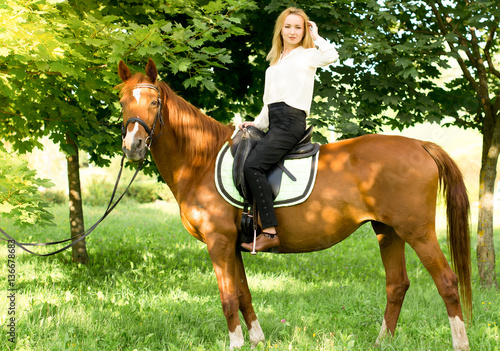Молодая девушка со светлыми волосами сидит верхом на коричневой лошади в лесу 