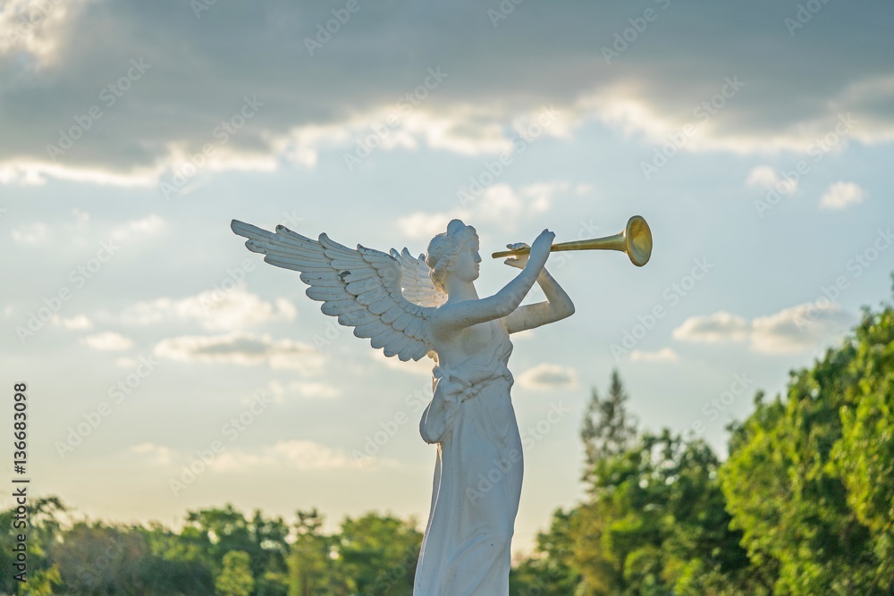 sculpture of angel blowing golden horn on sunrise sky clouds with trees ...