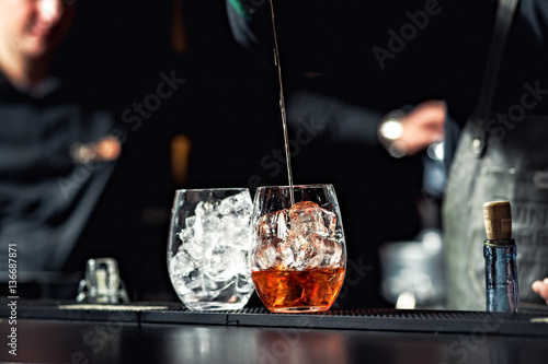 Closeup of bartender hands pouring alcoholic drink into a jigger to prepare a cocktail, with red bell pepper and peppercorn seeds in a serving glass.