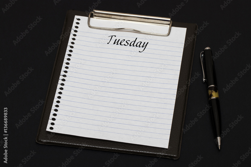 Top view of clipboard and white sheet written with Tuesday on bl Top view of clipboard and white sheet written with Tuesday on bl